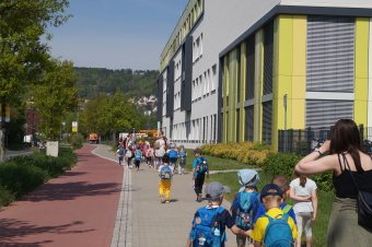 zu sehen sind Kinder auf einem Gehweg gehend, rechts steht ein großes Gebäude, links befindet sich die Straße