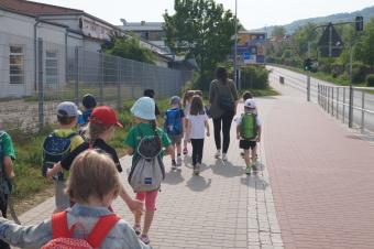 zu sehen sind Kinder, die mit einer Erzieherin eine STraße entlang gehen. Rechts ist ein Gebäude, links eine Straße