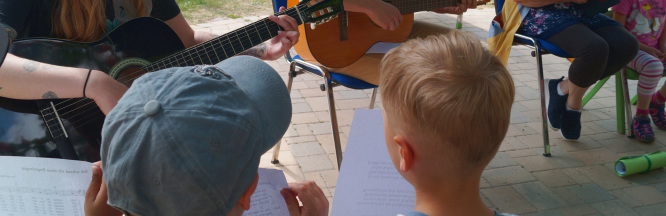 zu sehen sind Kinder mit weißen Blätter in der Hand, im Hintergrund sind 2 Gitarren zu erkennen, die wiederum durch 2 Erwachsene bespielt werden