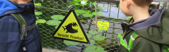 zu sehen sind zwei Kinder vor einem Teich. Zwischen ihnen ist ein gelbes Schild mit einem Fisch mit scharfen Zähnen und der Aufschrift "Vorsicht Piranhas"