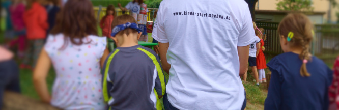 zu sehen sind mehrere Kinder und ein Erwachsener in Rückenansicht. Im Hintergrund sieht man verschwommen weitere Kinder