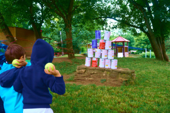 zu sehen sind Kinder in der Rückenansicht, die mit einem Ball auf einen Dosenturm werfen wollen