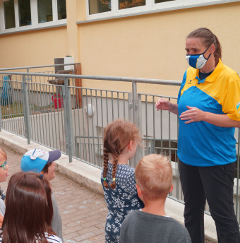 zu sehen sind Kinder, die einer Frau in gelb-blauer Kleidung zu hören