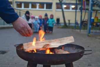 zu sehen ist eine Feuerschale in der Holz brennt, im Hintergrund sind Kinder. Im Vordergrund des Bildes wird ein Scheit Holz ins Feuer geworfen