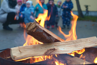 zu sehen ist ein Feuer im Vordergrund. Im Hintergrund stehen Kinder und schauen dem Feuer zu