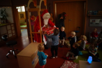 zu sehen ist der Weihnachtsmann an einem Rundbogen. Vor ihm sitzen viele Kinder. EIn Kind nimmt ein Geschenk entgegen.