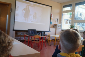 zu sehen sind Kinder, die auf einem Stuhl sitzen und auf eine Leinwand schauen. Auf dieser sind Personen zu sehen, die etwas zeigen.