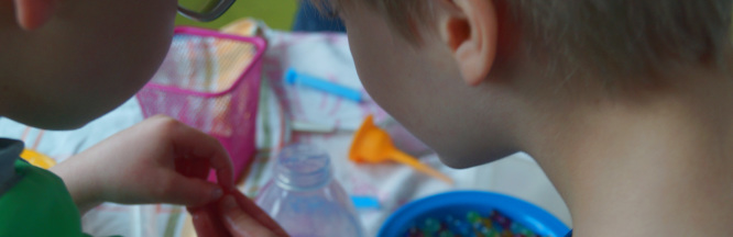 Zu sehen sind 2 Kinder, die sich etwas in der Hand des einen Kindes ansehen. Im Hintergrund ist eine blaue Schüssel, mit vielen bunten Kügelchen.