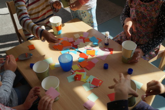 zu sehen sind Kinder an einem Tisch bastelnd. In der Mitte befinden sich viele bunte Papierschnipsel