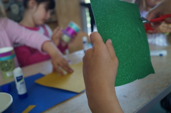 zu sehen ist ein Kind, dass mit einer Schere ein grünes Blatt Papier zerschneidet. Im Hintergrund bastelt ein weiteres Kind