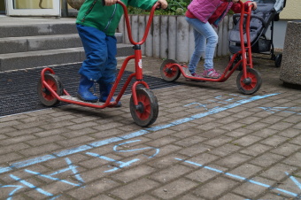 zu sehen sind zwei Kinder auf einem Roller. Vor ihnen auf dem Weg ist mit Kreide eine blaue Linie gezogen