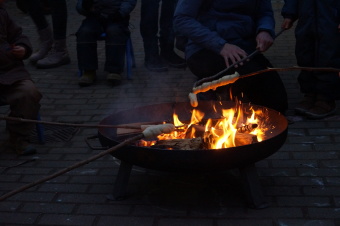 zu sehen ist ein Feuer in einer Feuerschale. Mehrere Personen halten Stöcke mit Teig am Ende in das Feuer