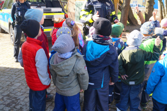 zu sehen sind Kinder vor einem Polizeiauto und einem Polizisten