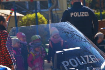 Zu sehen ist die Frontscheibe eines Polizeimotorrads, im Hintergrund steht ein Polizist mit Kindern.