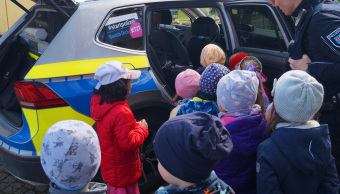 Zu sehen ist ein Polizeistreifenwagen mit offenen Türen. Die Kinder und ein Polizist schauen hinein.