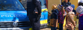 Zu sehen sind Kinder vor einem Polizeiauto. Zwei Polizisten stehen bei den Kindern.