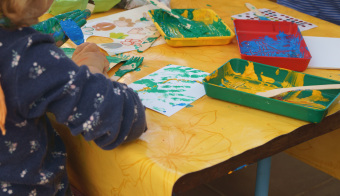 zu sehen ist eine angemalte Kinderhand, sowie Farbe und weiteres Bastelmaterial auf den Tisch