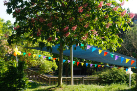 zu sehen ist ein kastanienbaum im Vordergrund. Im Hintergrund sind ein Sonnensegel und bunte Banner zu sehen