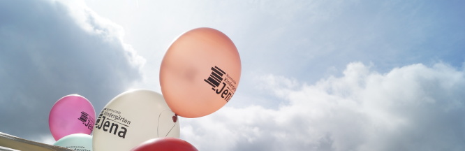 zu sehen sind bunte Luftballons mit der Aufschrift "kommunale Kindergärten Jena". Im Hintergrund sind aufbrechende Wolken mit durchscheinendem Sonnenlicht