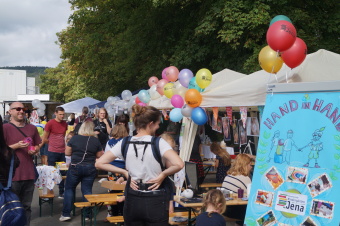 zu sehen ist eine Menschenmenge vor einem Stand mit bunten Luftballons.  Im Vordergrund ist ein Aufsteller mit der Aufschrift "Hand in Hand. Kommunale Kindergärten Jena" zu lesen