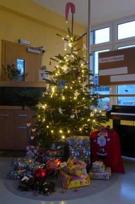 zu sehen ist das Foyer der Kita. Im Zentrum des Bildes ist ein geschmückter Weihnachtsbaum zu sehen, links ein Aquarium, rechts ein Klavier. Unter dem Weihnachtsbaum sind Geschenke zu sehen