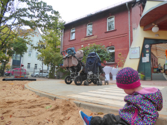 zu sehen ist ein Kind, welches vor einem Gebäude mit der Aufschrift: Fröbelhaus im Sandkasten sitzt