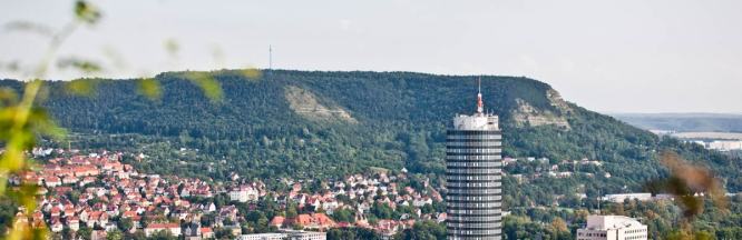 Zu sehen ist ein Weitblick auf Jena. Es sind verschiedene Häuser, sowie der JenTower zu erkennen. Im Hintergrund ist ein Berg zu sehen.