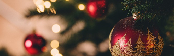 zu sehen ist eine rote Christbaumkugel, mit goldglitzerndem Weihnachtsbaummotiv. Im Hintergrund sind unscharf die Zweige eines Weihnachtsbaumes zu sehen