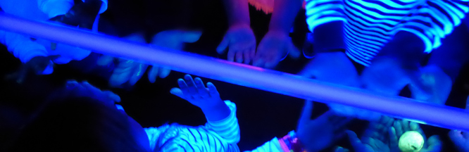 zu sehen sind Kinderhände unter einer Schwarzlichtlampe. Die Hände schillern in bunten Farben