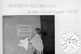 zu sehen ist ein schwarzweiß Bild auf dem eine Frau ein Tuch in der Hand hält. Vor ihr enthüllt sich eine Tafel an einer Hauswand mit der Aufschrift "Kinderkrippe Werner Selenbinder"