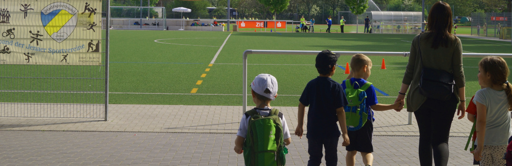 zu sehen sind Kinder mit einer Erzieherin, die auf eine grüne Wiese zu gehen.